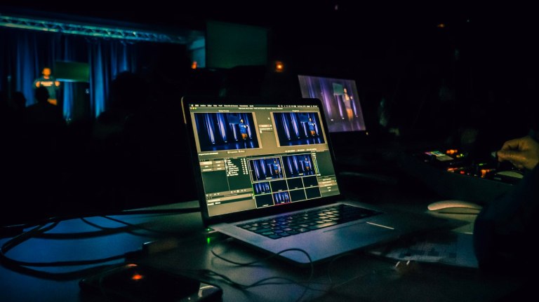 A dimly lit room with a laptop on a table displaying multiple video editing screens, labeled "Sample Post #3," and a person standing on stage in the background blurred in blue lighting.