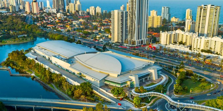 Aerial view of a large convention center surrounded by urban buildings and a water body, with a network of roads and green spaces harmoniously blending into the scene.