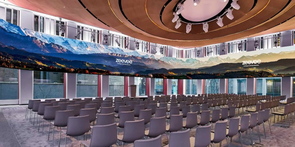 A modern conference room with rows of gray chairs facing a large, curved screen displaying a mountainous landscape with the word "zeevee" on it. Ceiling lighting fixtures are visible above.